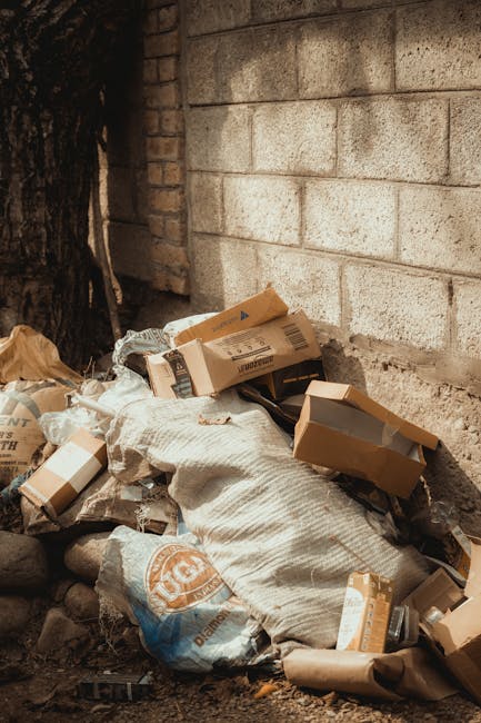 A pile of discarded cardboard boxes, some flattened and others partially intact, is situated against a weathered brick wall in an outdoor urban environment. The boxes vary in size, with some bearing printed labels and barcodes, and are made of brown corrugated material. Intermixed with the boxes is a white woven textile sack that appears somewhat torn and crumpled. Surrounding the pile are small rocks and dirt, indicating the scene is on a paved or dirt surface, possibly a back alley or side passageway. To the left, part of a tree trunk is visible, casting shadows over the rubbish and adding a natural element to the otherwise concrete and brick setting. The lighting is natural, subtly illuminating the scene, and the overall impression is of a cluttered area awaiting collection or clearance. This visual relates to independent waste handling or the disposal of household or office rubbish, consistent with the services provided by Waste Clearance Bermondsey for efficient rubbish removal in the Bermondsey area.