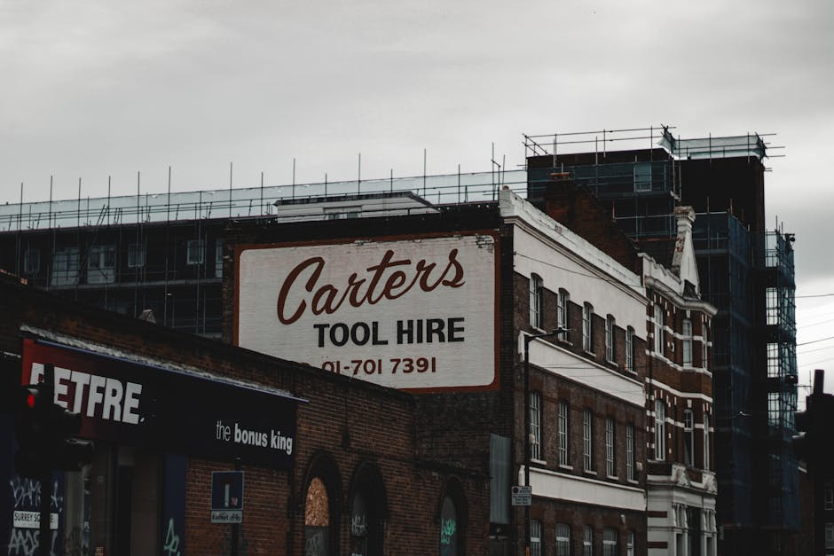 The image depicts a multi-story brick building under construction or renovation, with scaffolding erected around the upper floors and on the sides. On the building's façade, there is a prominent white sign with red and black lettering for 'Carter's Tool Hire,' including a contact phone number. The lower part of the building features large windows and arched doorways, some partially obscured by a dark awning that advertises 'the bonus king.' The overall scene is set in an urban environment, with overcast skies casting diffuse lighting over the scene. The presence of scaffolding and signage suggests ongoing building work or refurbishment, consistent with private or independent construction-related activities, often requiring rubbish clearance services by companies like Waste Clearance Bermondsey, who handle waste generated during such projects.