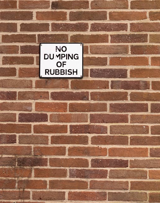 A close-up view of a brick wall with a rectangular, white metal sign mounted on it, displaying the message 'NO DUMPING OF RUBBISH' in bold black letters. The bricks are in various shades of red, brown, and orange, laid in a horizontal pattern with visible mortar joints. The sign is positioned slightly above the center of the image, and the lighting appears natural, casting subtle shadows on the wall. This setting exemplifies a common site where restrictions on rubbish disposal are enforced, aligning with themes of waste management and private rubbish clearance services such as those offered by Waste Clearance Bermondsey.